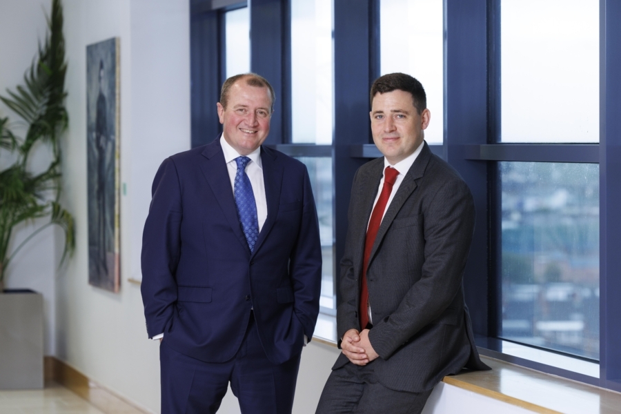 Two smiling men in business suits, shirts and ties against a window with a city backdrop